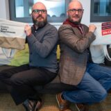 Finn und Jan Siebert sitzen Rücken an Rücken auf einer Couch, halten Taschen ihrer jeweiligen Partei in die Höhe und schielen sich argwöhnisch an.