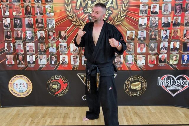 Chris Stallmann steht vor einer Wand mit der Aufschrift „MAA“. Das steht für Martial Arts