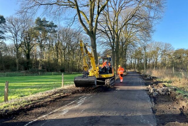 Ein gelber Bagger steht auf einem Wirtschaftsweg. Die Seitenstreifen sind des Weges sind aufgerissen. Ein Mann in orangefarbener Arbeitsmontur steht hinter dem Bagger mit einer Schaufel. Im Fahrerhaus des Baggers sitzt ein weiterer Bauarbeiter. Im Hintergrund ist ein Wäldchen zu sehen. Der Himmel ist blau und wolkenlos.