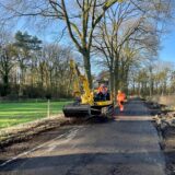 Ein gelber Bagger steht auf einem Wirtschaftsweg. Die Seitenstreifen sind des Weges sind aufgerissen. Ein Mann in orangefarbener Arbeitsmontur steht hinter dem Bagger mit einer Schaufel. Im Fahrerhaus des Baggers sitzt ein weiterer Bauarbeiter. Im Hintergrund ist ein Wäldchen zu sehen. Der Himmel ist blau und wolkenlos.
