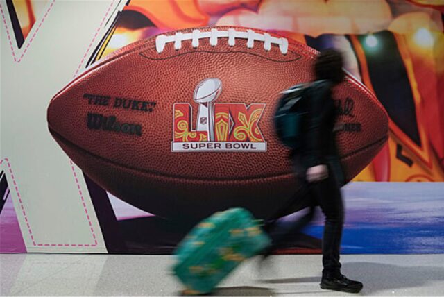 Eine Person mit Reisekoffer geht an einem Football in Menschengröße vorbei. Auf dem Football steht Super Bowl.