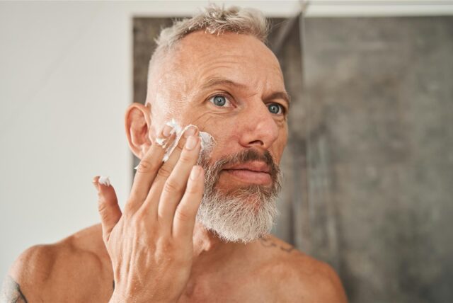 Man mit grauem Haar und Bart trägt eine weiße Creme auf seine rechte Wange auf und schaut dabei in den Spiegel.
