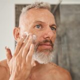 Man mit grauem Haar und Bart trägt eine weiße Creme auf seine rechte Wange auf und schaut dabei in den Spiegel.