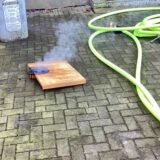 Auf einer gepflasterten Fläche liegt ein qualmendes Holzbrett, daneben ein hellgrüner Wasserschlauch.