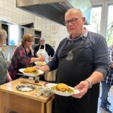 Mehrere Personen in einer Küche, ein Mann hat zwei fertig angerichtete Teller mit Essen in der Hand.