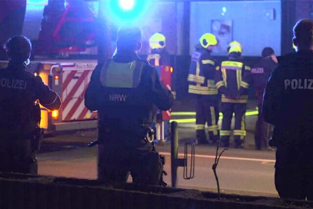 Polizisten mit dem Rücken zur Kamera, im Hintergrund Feuerwehrkräfte an einem Löschfahrzeug.