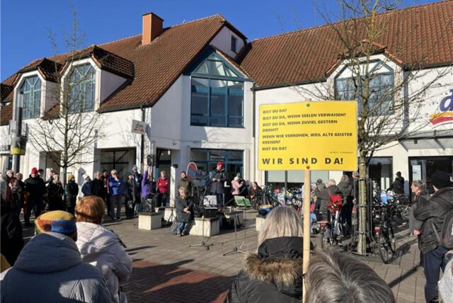Menschen am Mikrofon vor einer Menschenmenge, ein Schild mit dem Aufdruck „Wir sind da“.