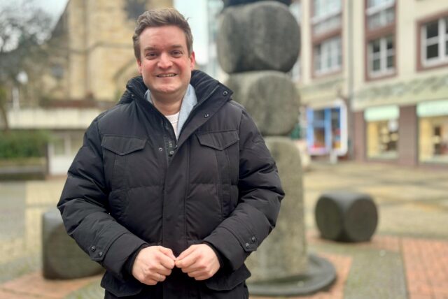 Michael Breilmann steht lächelnd am Lambertusbrunnen. Im Hintergrund ist die Lambertuskirche zu erahnen.
