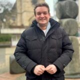 Michael Breilmann steht lächelnd am Lambertusbrunnen. Im Hintergrund ist die Lambertuskirche zu erahnen.