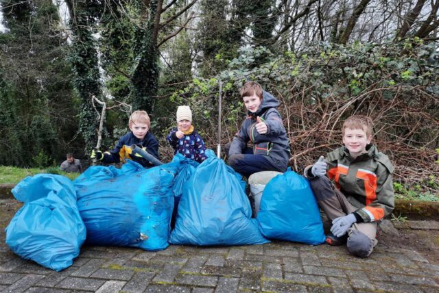 Vier Kinder stehen hinter vier großen blauen Säcken, die mit Müll gefüllt sind und haben Handschuhe sowie eine Müllzange in der Hand