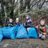 Vier Kinder stehen hinter vier großen blauen Säcken, die mit Müll gefüllt sind und haben Handschuhe sowie eine Müllzange in der Hand
