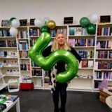 Britta Spengler steht mit einer grünen Luftballon-Fünf vor Regalen in ihrer Buchhandlung in Recklinghausen-Süd.