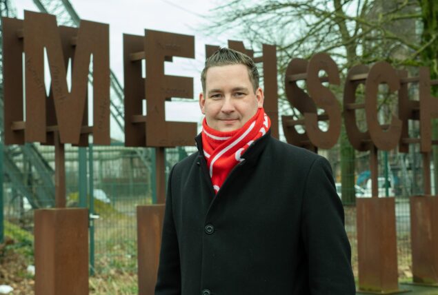 Brian Nickholz, SPD-Bundestagsabgeordneter, steht vor einem Schriftzug aus Bronze, der das Wort „Mensch“ bildet, neben der La Victoria Eventhalle auf dem ehemaligen Schachtgelände AV 1/2 in Marl.