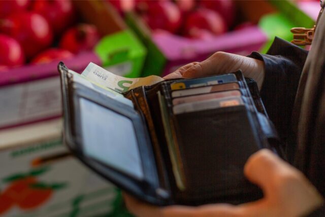 Eine Hand hält ein Portemonnaie, im Hintergrund liegt Obst in einer Supermarkt-Auslage