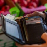Eine Hand hält ein Portemonnaie, im Hintergrund liegt Obst in einer Supermarkt-Auslage