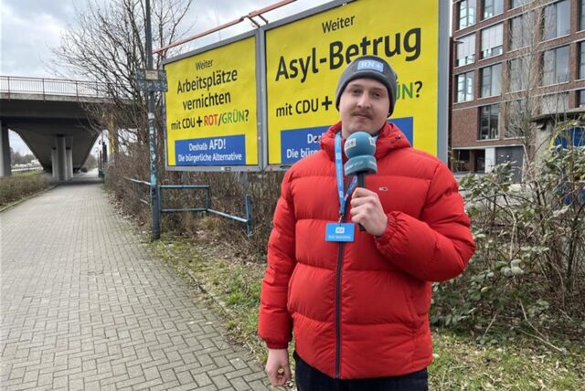 Reporter Tim Hübbertz steht vor zwei gelben Wahlplakaten mit Slogans für die AfD.
