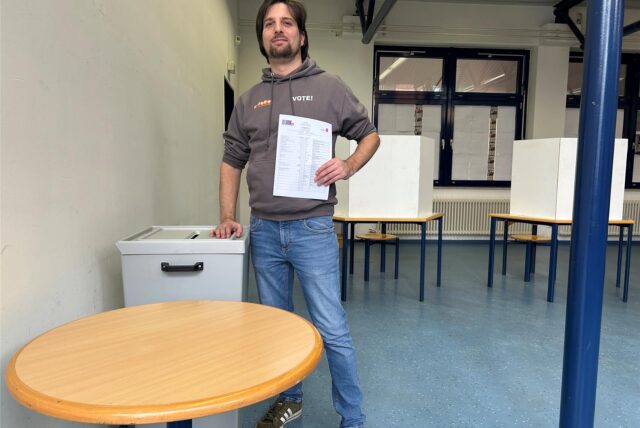 Ein Mann steht neben einer Wahlurne in einem Raum an der Gesamtschule in Waltrop. In seiner linken Hand hält er einen Wahlzettel und im Hintergrund sind Wahlkabinen aufgebaut.