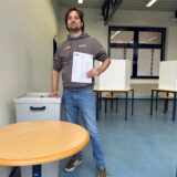 Ein Mann steht neben einer Wahlurne in einem Raum an der Gesamtschule in Waltrop. In seiner linken Hand hält er einen Wahlzettel und im Hintergrund sind Wahlkabinen aufgebaut.