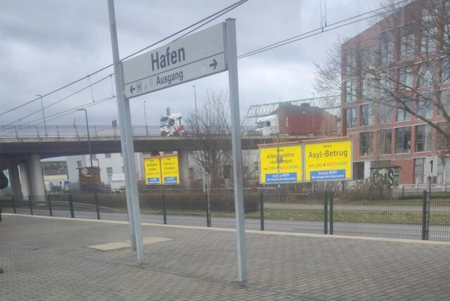 Blick aus der Stadtbahn auf die Haltestelle Hafen, hinter dem Haltestellenschild leuchtend gelbe Plakate mit großen schwarzen Aufschriften.