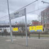 Blick aus der Stadtbahn auf die Haltestelle Hafen, hinter dem Haltestellenschild leuchtend gelbe Plakate mit großen schwarzen Aufschriften.