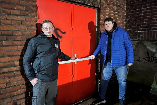 Lukas Boeckenholt, Geschäftsführer von Schacht3Events (r.), und Marco Knopp (Stellvertreter) stehen auf dem Zechengelände Schlägel&Eisen in Herten-Langenbochum vor der Halle mit roter, verschlossener Stahltür und fettem Stahlriegel davor.