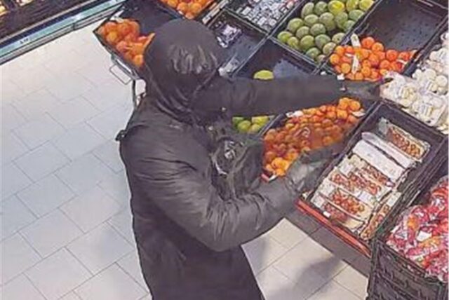 Ein schwarz gekleideter und vermummter Mann steht vor dem Obstregal eines Supermarktes.
