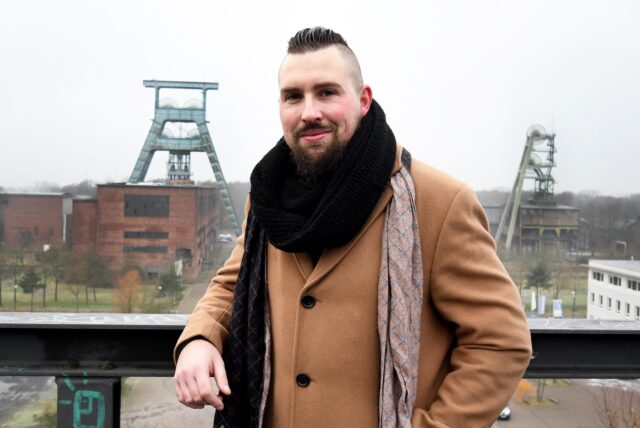 Der in Herten geborene Rapper Sebastian Czyszczon steht auf einem Balkon der Halde Hoheward und lehnt sich an ein Geländer. Im Hintergrund sind Fördertürme und Gebäude der Zeche Ewald zu sehen.