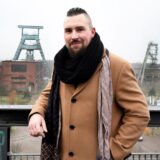 Der in Herten geborene Rapper Sebastian Czyszczon steht auf einem Balkon der Halde Hoheward und lehnt sich an ein Geländer. Im Hintergrund sind Fördertürme und Gebäude der Zeche Ewald zu sehen.