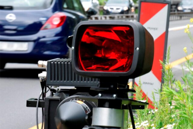 Ein mobiles geschwindigkeitsmessgerät steht am Straßenrand. Im Hintergrund ist der Seitenstreifen, sowie ein Auto dass von der Kamera wegfährt zu sehen. Weitere Autos kommen aus der entgegengesetzten Fahrtrichtung.