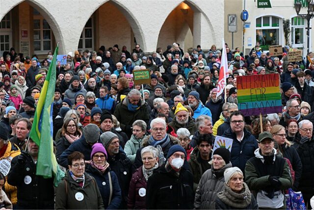 Hunderte Menschen stehen auf einem Marktplatz vor einem weißen Gebäude und halten Schilder und Fahnen hoch
