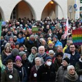 Hunderte Menschen stehen auf einem Marktplatz vor einem weißen Gebäude und halten Schilder und Fahnen hoch