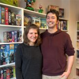 Jennifer und Johannes stehen vor einem großen Regal voll mit verschiedenen Spielen.