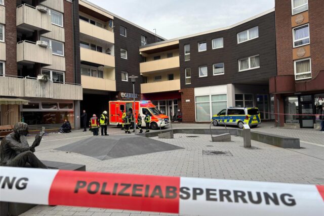 Auf dem Place d’Arras in der Hertener Innenstadt stehen Einsatzkräfte und Fahrzeuge der Polizei und der Feuerwehr. Im Bildvordergrund ist ein rot-weißes Flatterband mit dem Aufdruck Polizeiabsperrung zu sehen.