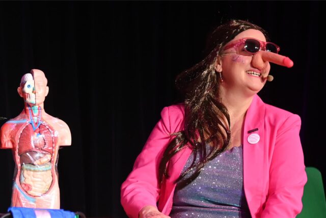 Eine Frau mit pinkfarbenem Blazer und einem Plastikpenis auf der Nase.