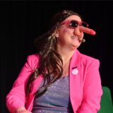 Eine Frau mit pinkfarbenem Blazer und einem Plastikpenis auf der Nase.