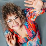 Die Dortmunderin Ulrike (41) lächelt in die Kamera. Sie hat gelocktes Haar, trägt eine Brille und ein buntes Kleid. Sie nimmt an der neuen Staffel des Reality-Formats „Big Brother“ teil.