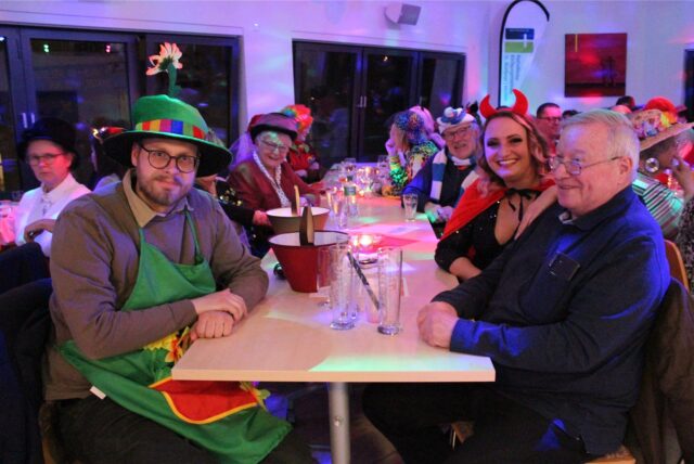 Kostümierte Menschen sitzen an einem Tisch und blicken in die Kamera.