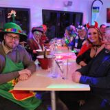 Kostümierte Menschen sitzen an einem Tisch und blicken in die Kamera.