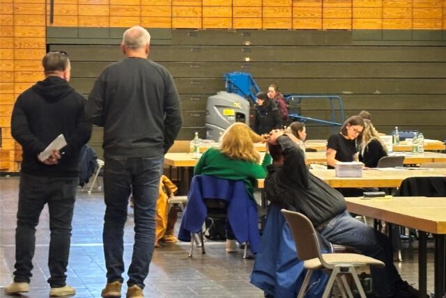 Bundestagswahl 2025 in Castrop-Rauxel: In der Europahalle werden die Stimmzettel aus der Briefwahl gezählt. Zwei Männer schlendern zwischen den Tischen hindurch und beobachten die Vorgänge.