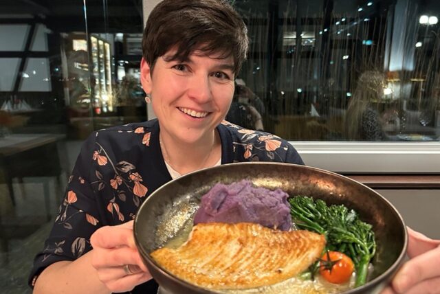 Jessica Will hält einen Teller mit ihrem Hauptgericht in die Kamera: Steinbuttfilet mit lilafarbenem Kartoffelstampf, Brokkoli und Tomaten.