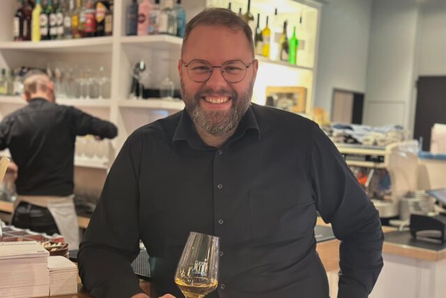 Ein Mann mit Brille hält ein Weinglas in der Hand und lacht in die Kamera.