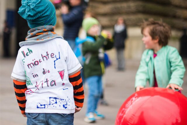 Kinder spielen, eins trägt ein T-Shirt mit der Aufschrift „Ich möchte in die Kita“.