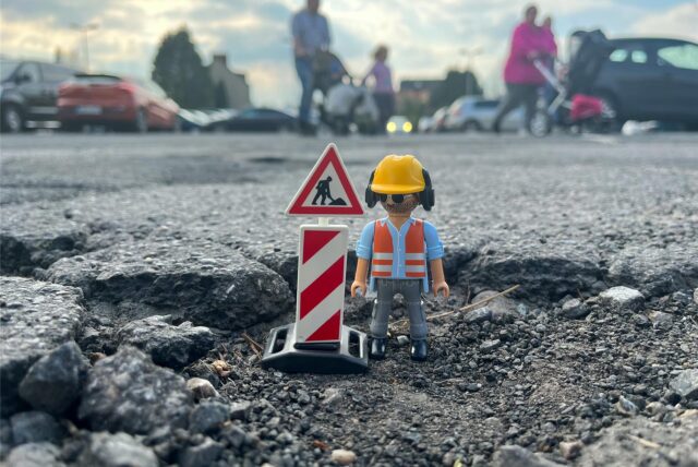 Eine Playmobil-Figur eines Bauarbeiters steht in einem Schlagloch.