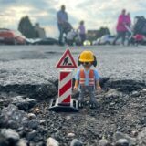 Eine Playmobil-Figur eines Bauarbeiters steht in einem Schlagloch.