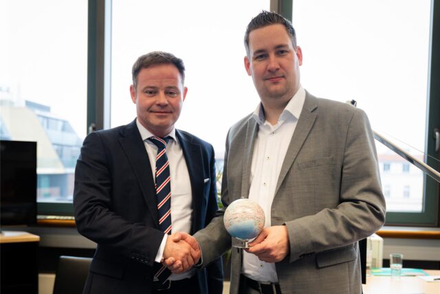 Bundestagsabgeordnete stehen in einem Büro und geben sich die Hand. Links der neue CDU-Bundestagsabgeordnete Lars Ehm, rechts der ausscheidende SPD-Bundestagsabgeordnete Brian Nickholz, der einen kleinen Globus in der linken Hand hält.