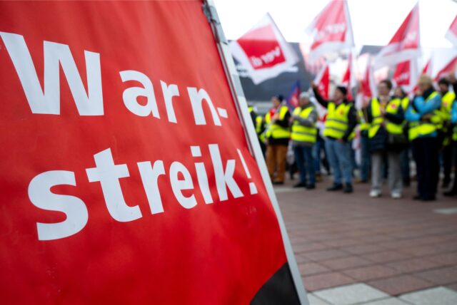 Ein Plakat mit der Aufschrift „Warnstreik“. Im Hintergrund sind Streikende zu sehen.