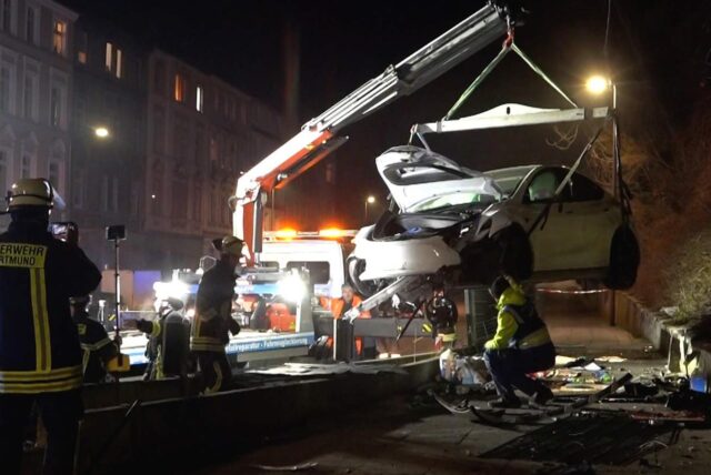 Ein weißer, deutlich demolierter Tesla hängt an der Aufhängung eines Kranwagens. Man erkennt ein Trümmerfeld am Fahrbahnrand und auf dem Bürgersteig.