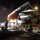 Ein weißer, deutlich demolierter Tesla hängt an der Aufhängung eines Kranwagens. Man erkennt ein Trümmerfeld am Fahrbahnrand und auf dem Bürgersteig.
