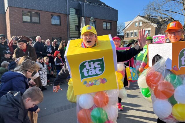 Eine Frau im Tic-Tac-Kostüm winkt in die Kamera, viele Menschen gucken sich die Kinderkarnevalsparade an.