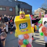 Eine Frau im Tic-Tac-Kostüm winkt in die Kamera, viele Menschen gucken sich die Kinderkarnevalsparade an.